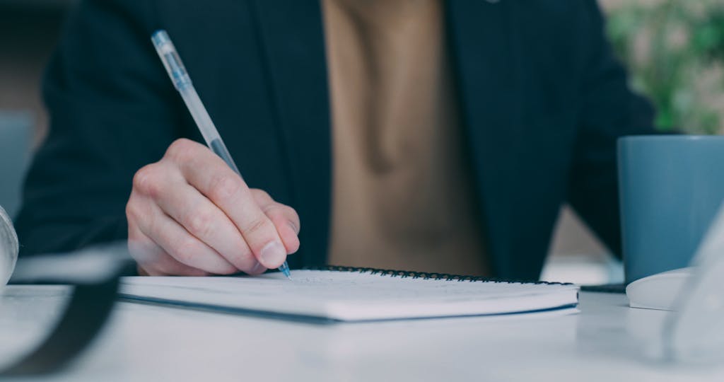 A person writing notes with a pen on a spiral notebook, indicating work or study.